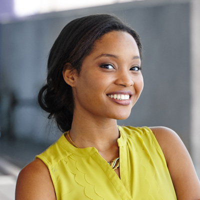 A young woman with a radiant smile, wearing a yellow top and posing against a backdrop of a building.